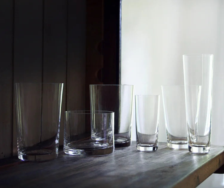 Six clear crystal deborah ehrlich drinking glasses in various shapes and sizes arranged on a dark wooden shelf against a softly lit indoor background.