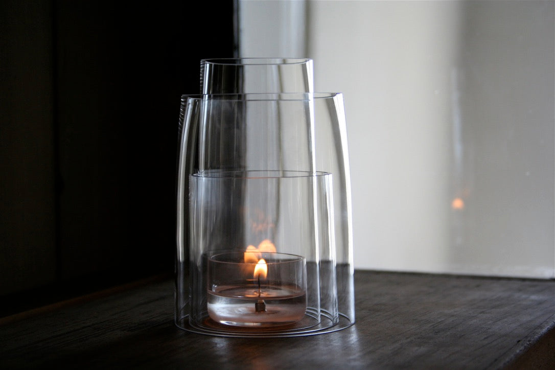 a clear tea light candle lit inside three nesting hand blown pieces of crystal on a dark wood shelf in front of a light window