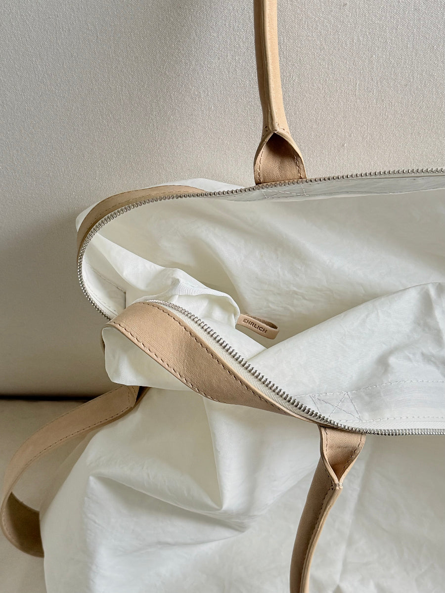 Close-up of a soft white sail fabric bag with beige leather handles and trim, showing the interior lining and zipper.