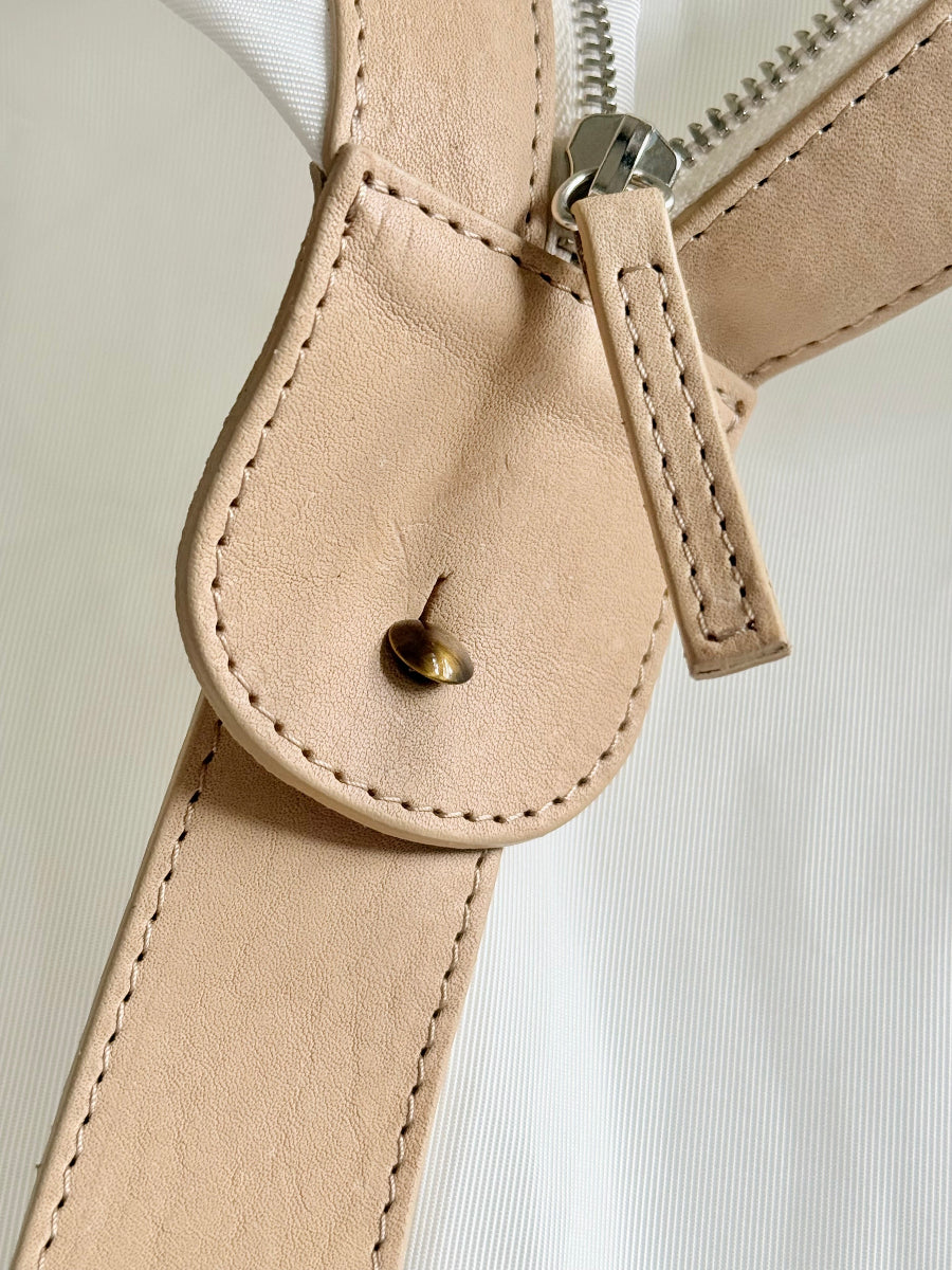 Close-up of a beige leather strap with a silver metal zipper pull and a small brass button, attached to a white ribbed sailcloth travel bag.