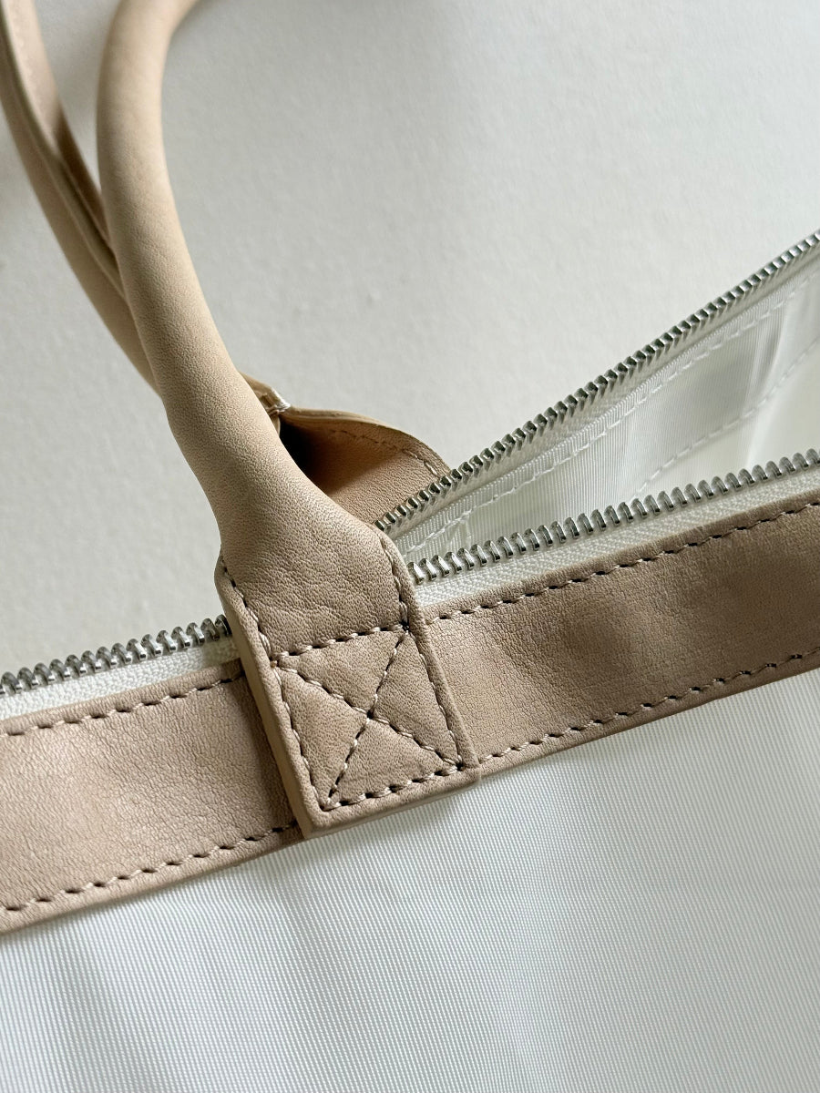 Close-up of a beige leather handle attached to a white travel bag with a silver zipper.