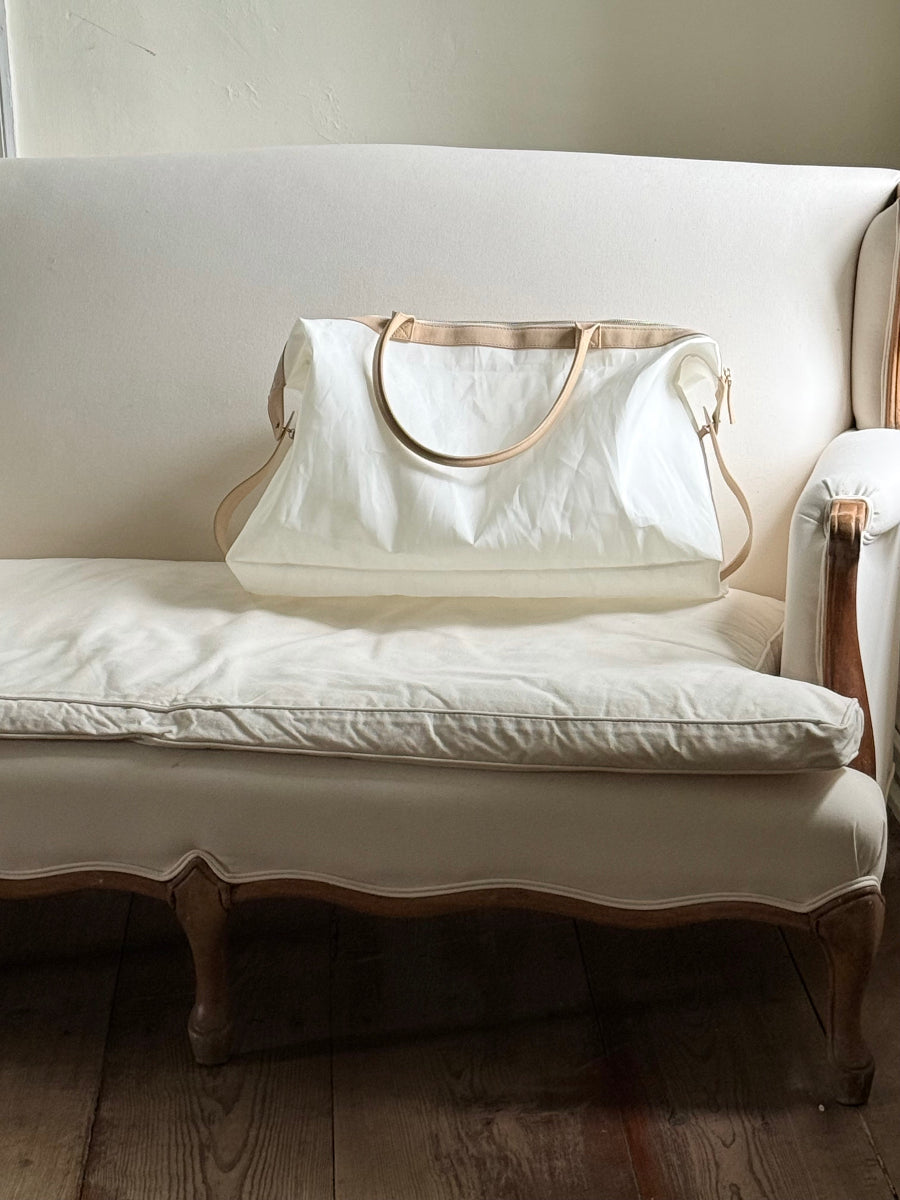 A large off-white sailcloth travel bag with tan leather handles rests on a matching off-white upholstered sofa in a softly lit interior with wooden flooring.