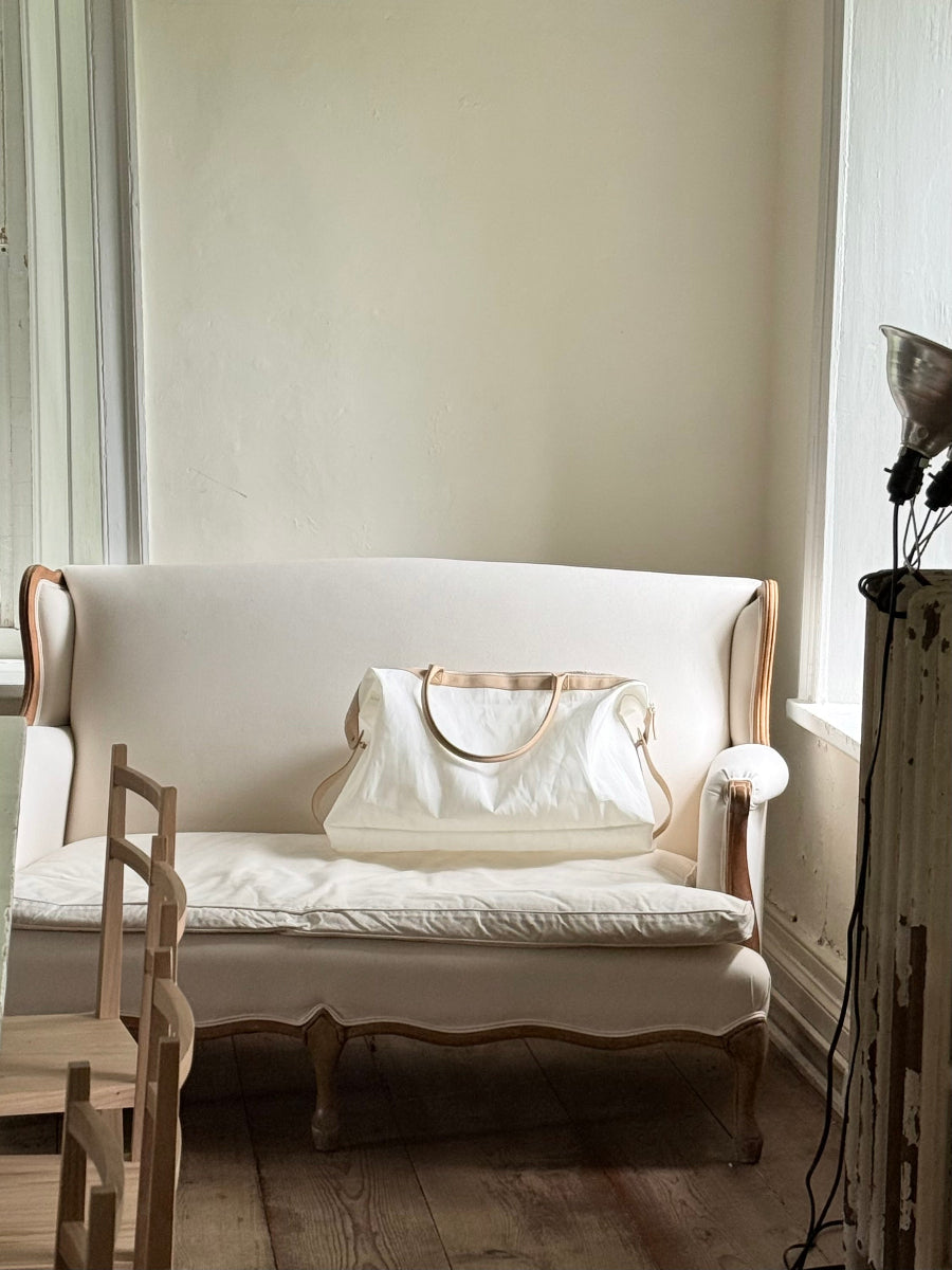 A cream-colored sailcloth travel bag with tan leather handles rests on a cream wingback sofa in a softly lit room with light wood floors and neutral walls.
