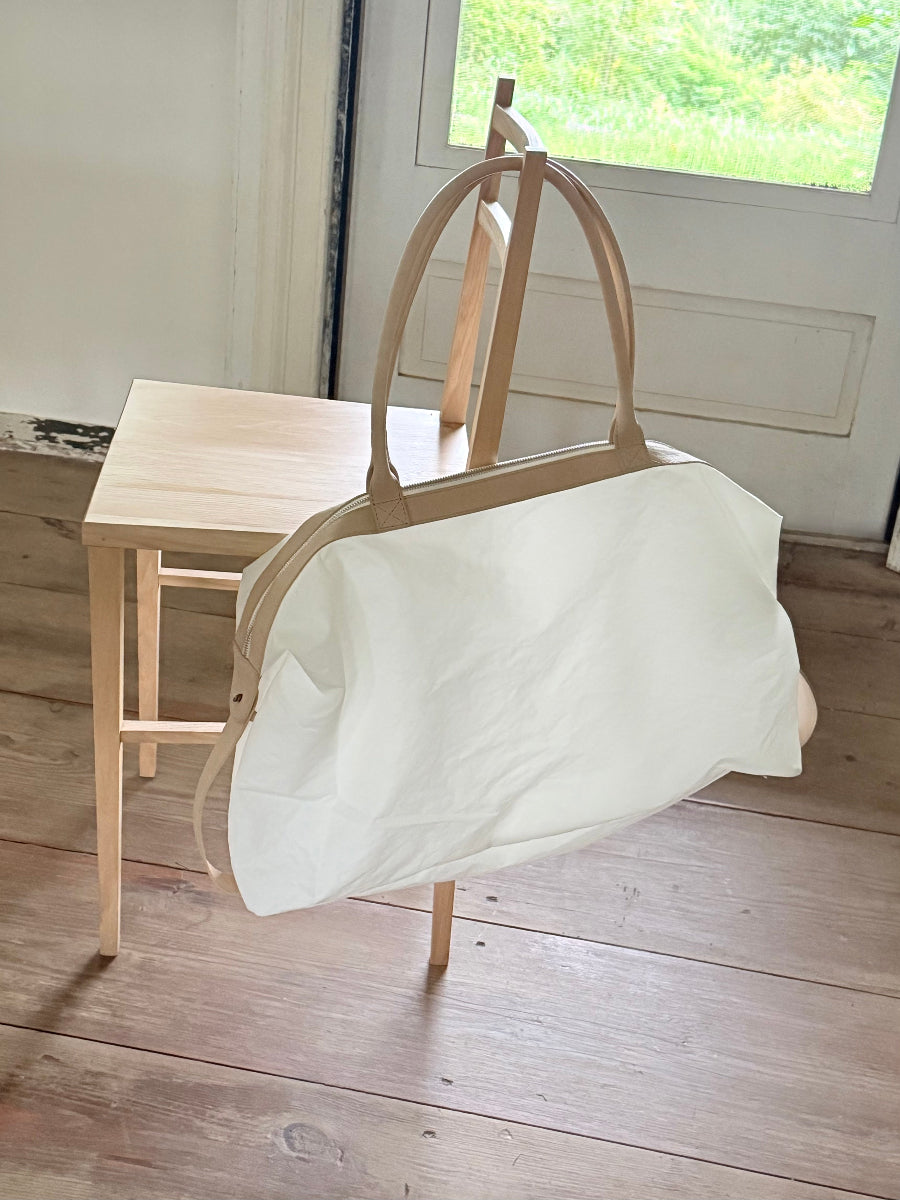 A large white travel bag with light tan leather handles and trim, draped over a light wood chair in a bright room with a wooden floor and window.