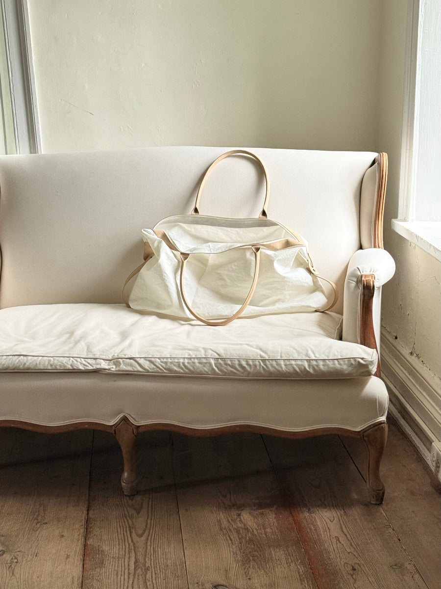A cream sailcloth travel bag with tan leather handles rests on an off-white upholstered wooden sofa in a softly lit room with wood floors.