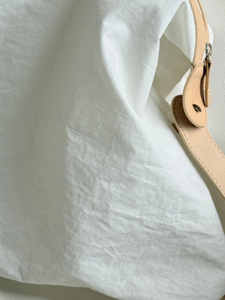 Close-up of a white crinkled sailcloth travel bag with a light beige leather strap and silver metal hardware.