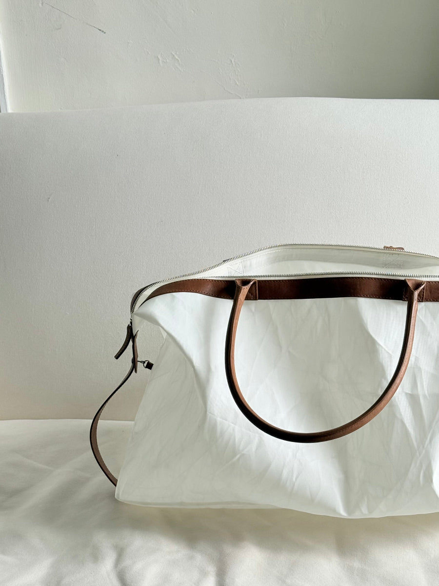 A white sailcloth travel bag with brown leather handles and trim, resting on a white fabric surface against a white background.