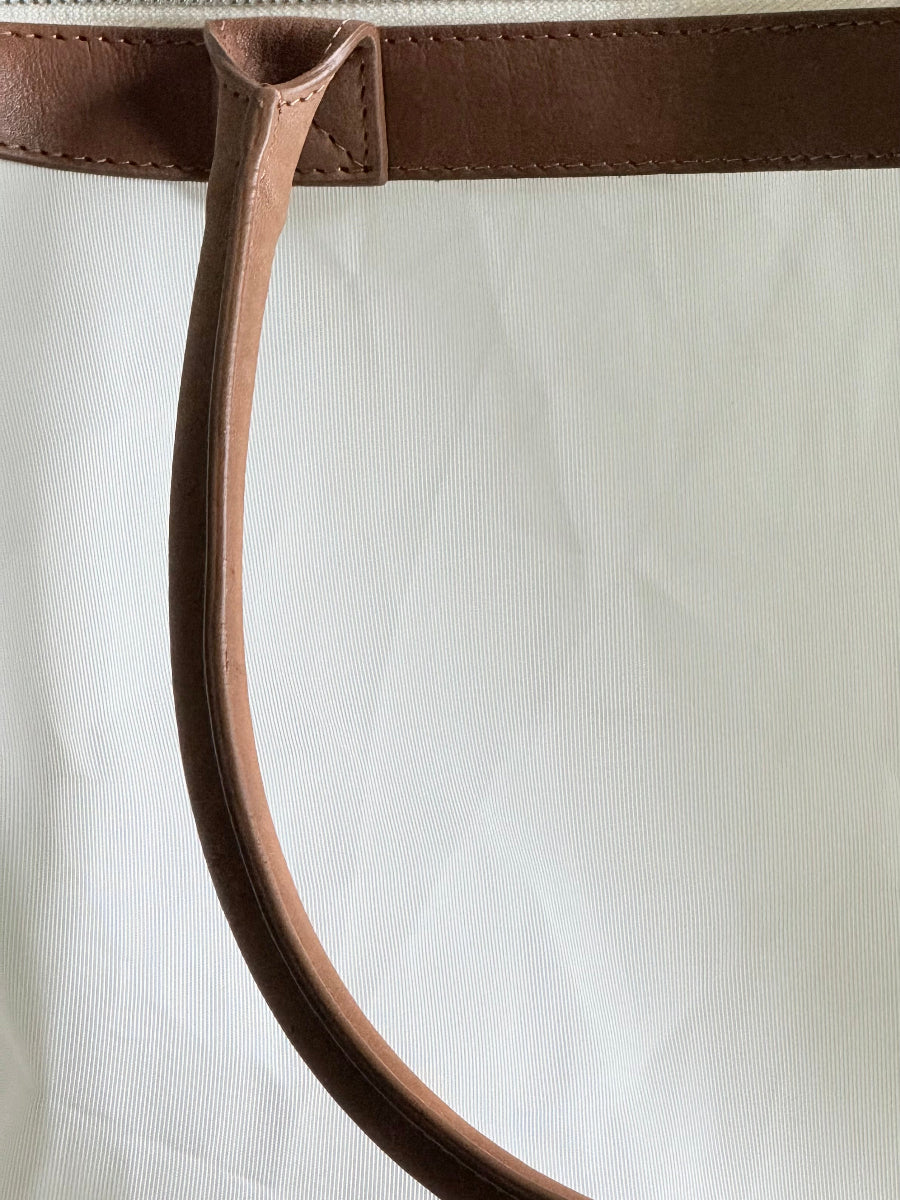 Close-up of a white sailcloth travel bag with a brown leather handle attachment and a matching brown leather top trim.