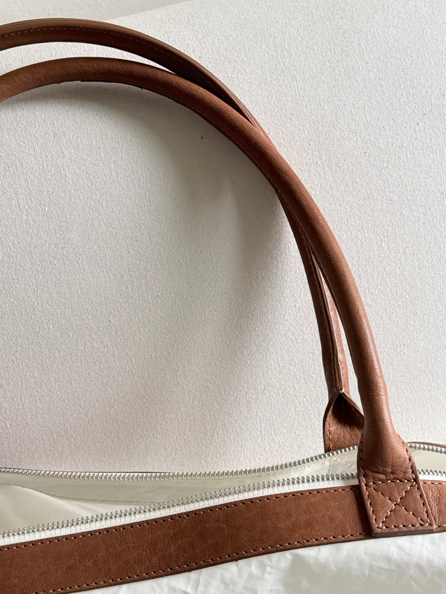 Close-up of a brown leather travel bag handle attached to a white sailcloth travel bag with a silver zipper, set against a plain off-white background.
