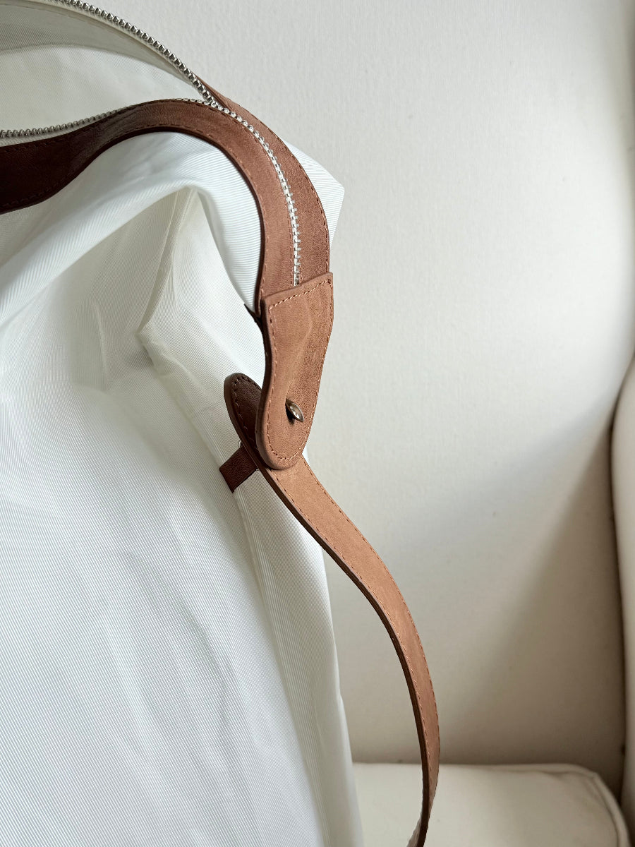 Close-up of a light brown leather strap with a silver-toned metal zipper, and cast brass fastener, attached to a white sail travel bag against a neutral cream background.