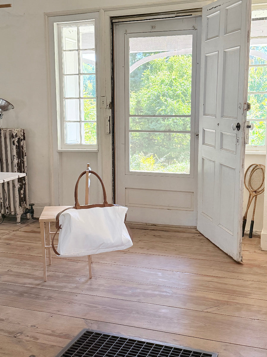 A white sail travel bag with brown leather handles hangs from a wooden chair in a bright, sunlit room with light wood floors and an open white screen door behind it.