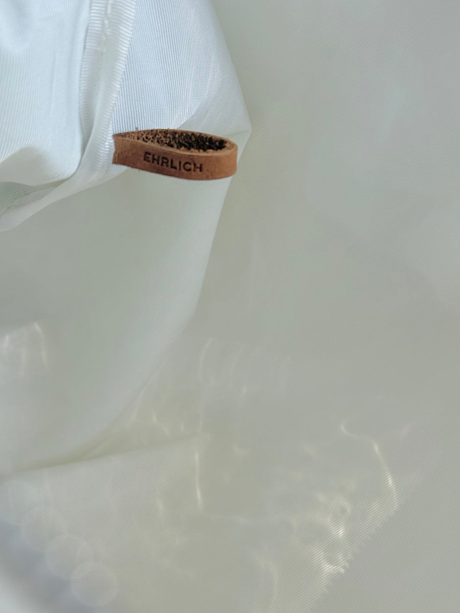 A close-up of a white sail cloth with a small tan leather tab reading EHRLICH, photographed against a soft white background with dappled light.