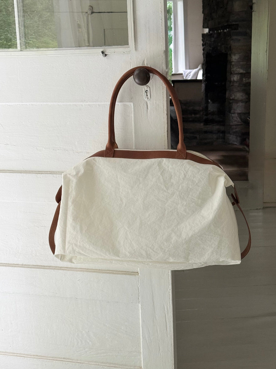 An off-white crinkled sail travel bag with brown leather handles and trim, hanging on a white wooden door in a home interior with s stone fireplace in the background.