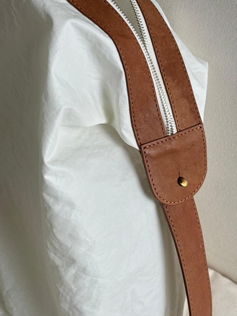 A close-up of a brown leather zipper strap with a silver zipper and cast brass metal button, draped over a white fabric surface.