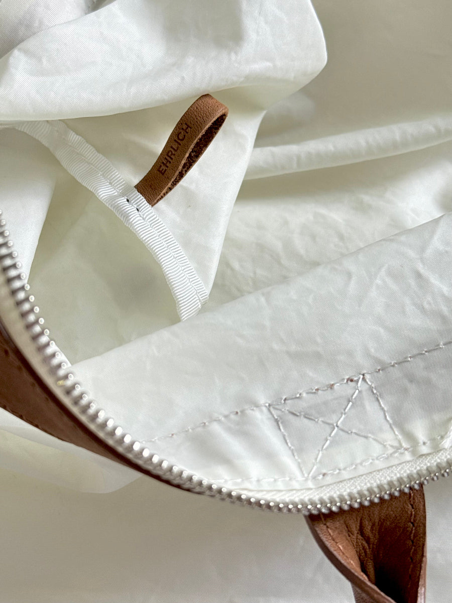 Close-up detail of a white fabric travel bag with a brown leather zipper pull and a small brown brand tab reading EHRLICH.