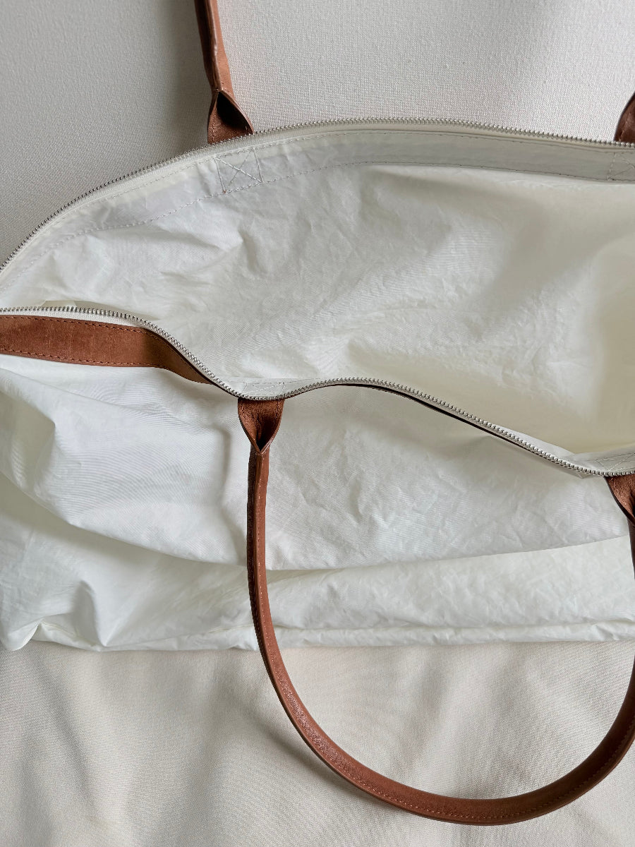 Close-up of a white fabric travel bag with brown leather handles and trim, photographed against a light neutral background.
