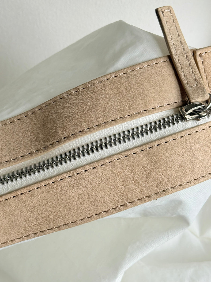 Close-up of a beige leather zipper with a silver metal zipper and white interior lining, resting on a white fabric background.