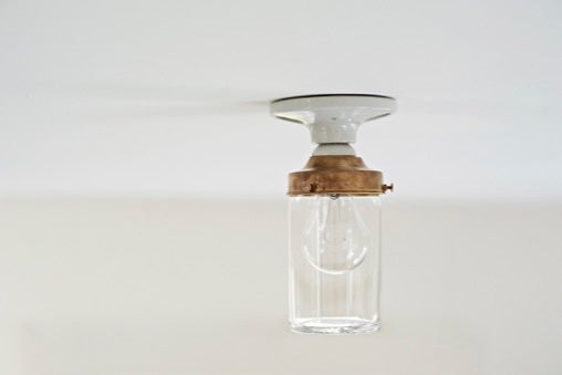 A minimalist ceiling light fixture with a clear crystal glass jar shade, a warm brass metal collar, and a white porcelain ceiling mount, shown against a plain white ceiling.