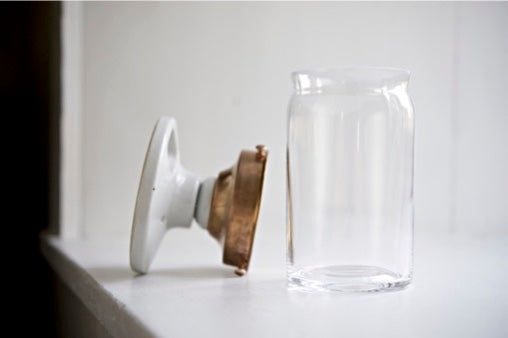 A clear glass jar with a white screw-on lid leaning against a white surface, with a copper-toned metal band around the neck, photographed in a bright indoor setting.