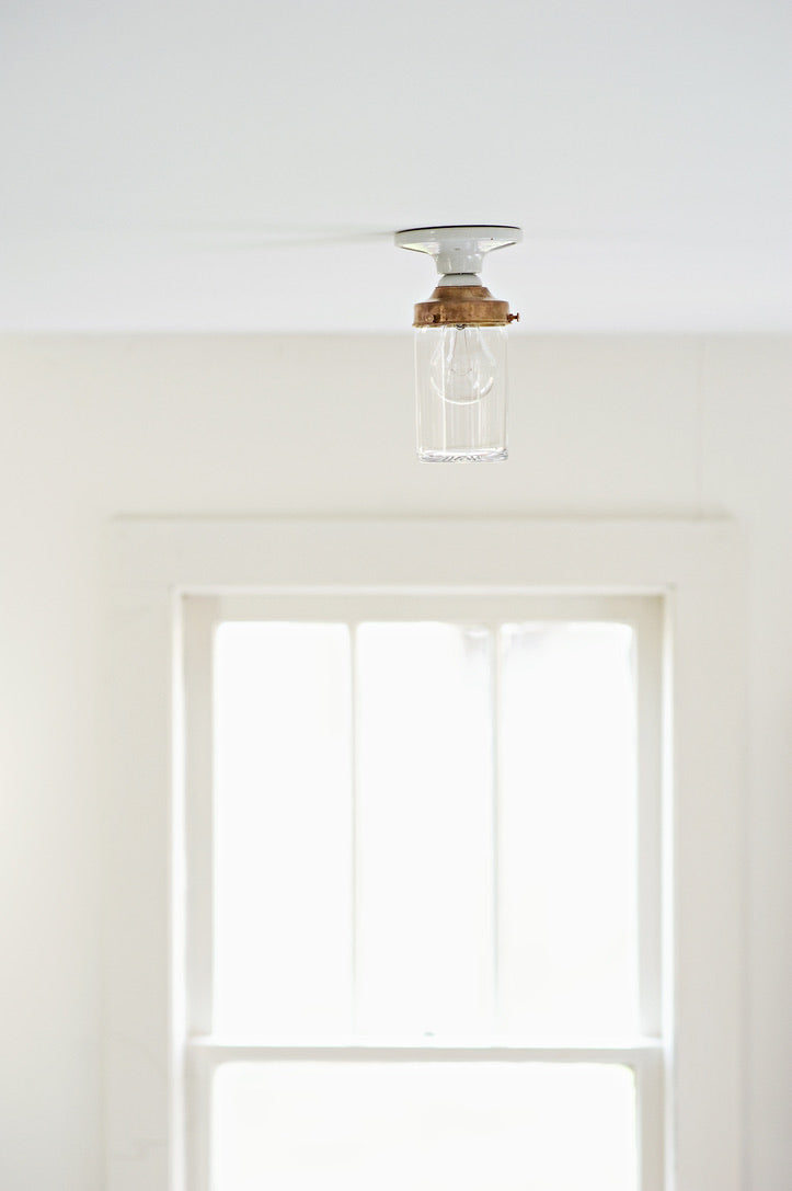A minimalist porcelain ceiling light fixture with a clear crystal glass jar shade and a copper colored brass collar, mounted on a white ceiling above a bright window.