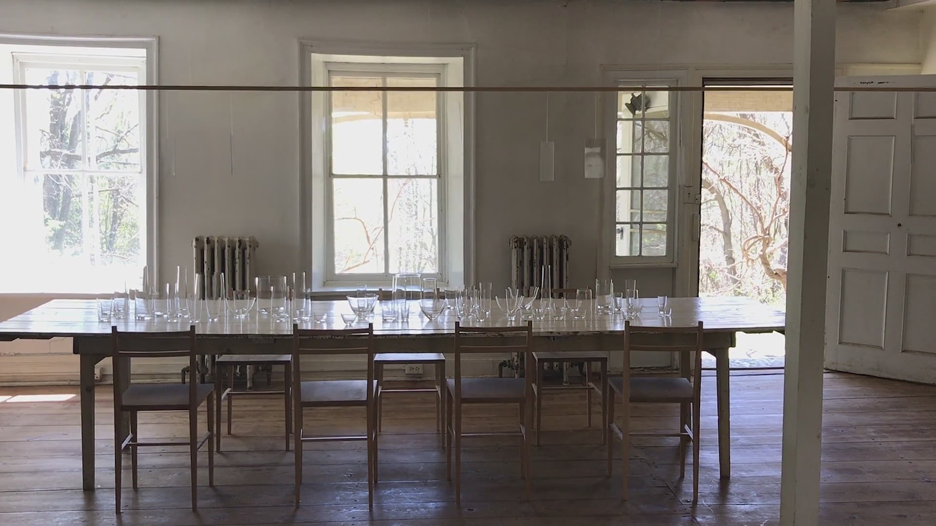 long white farmhouse table with wooden chairs around it, with crystal glassware by deborah ehrlich scattered on the table, in an historic house with wooden wide plank floors and tall windowpanes with deep sills, and a light mobile moving in the wind reflecting light.