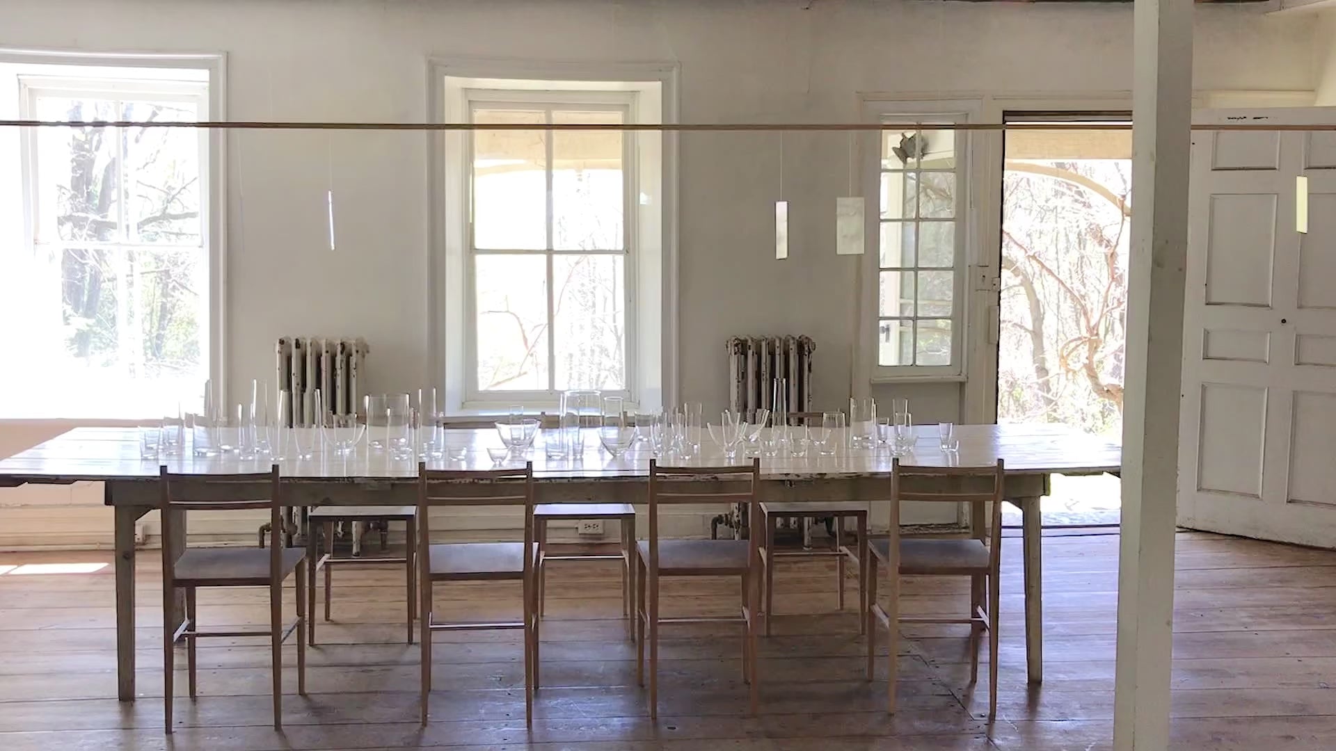 long white farmhouse table with wooden chairs around it, with crystal glassware by deborah ehrlich scattered on the table, in an historic house with wooden wide plank floors and tall windowpanes with deep sills, and a light mobile moving in the wind reflecting light.